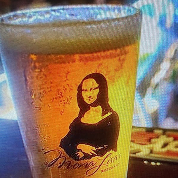A Mona Lisas Ristorante beer glass with the Mona Lisa logo and condensation, with a pizza in the background.